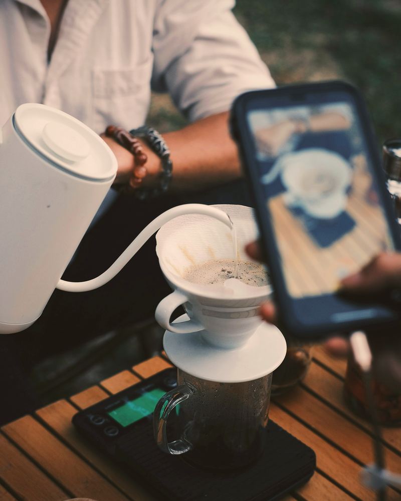 A hand holds a phone up to film coffee being poured from a kettle into a pour-over dripper on a wooden café table.