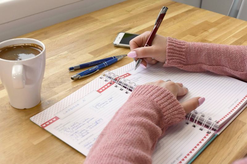 Hands writing in a planner on a wooden table beside a mug and a phone.