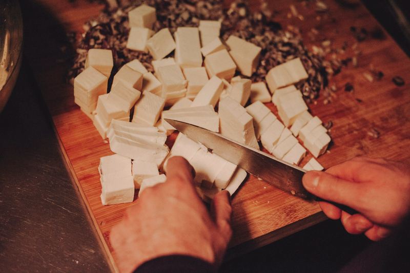 A hand cutting cubes of cheese on a wooden board with a long knife.