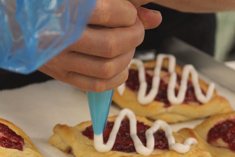 A hand piping white icing onto jam pastries on a tray.